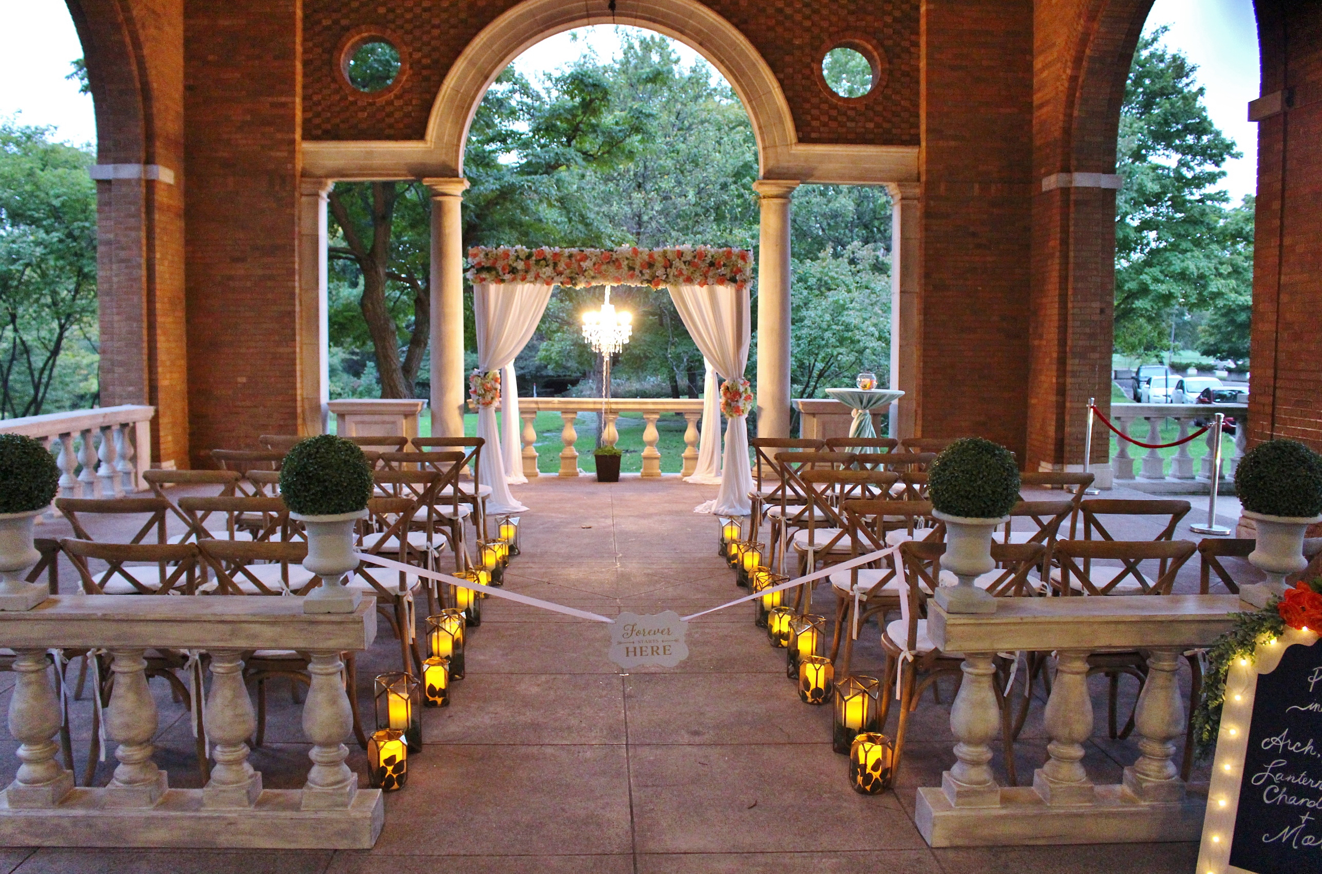 Outdoor wedding venue decorated with lights, flowers, and white fabric draping. Wooden cross-back chairs are lined up for guests.